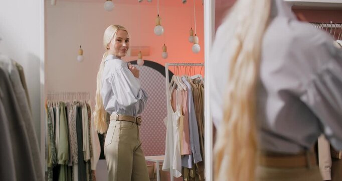 Stylish blonde woman looking at her reflection and adjusting shirt collar while trying on an outfit in a fashion boutique. Young female shopper stands in front of mirror in a soft pastel retail