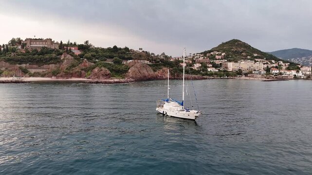 Sailboat Drifting Near Coastal Village. Singlemasted Yacht Glides On Glassy Water Past Hillside Homes Dusk Light, Gentle Swell And Mooring Buoys Evoke Serene Sailing And Leisure Travel.
