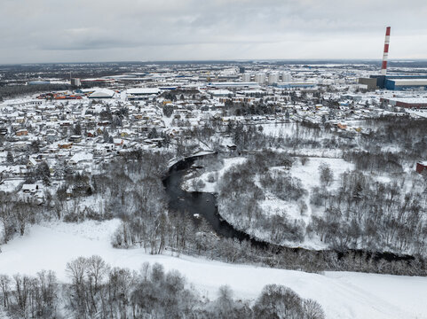 Aerial view of Tallinn winter cityscape with snow covered houses, winding river and frozen landscape under cloudy sky. Industrial district with Iru Power Plant chimney in distance, cold Baltic weather