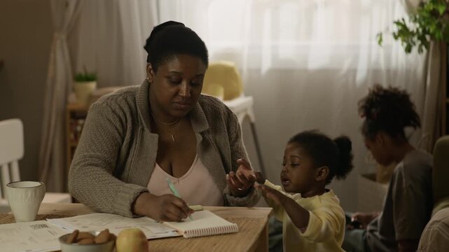 Handheld medium shot of young African American mother placing crying toddler girl on laps while reviewing bills and managing family budget at table, with her teenage daughter using phone in background