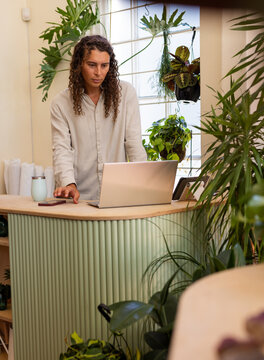 Working non-binary owner wearing cream shirt, typing on laptop at plant counter with smartphone