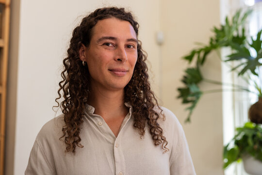 Standing nonbinary adult wearing cream buttonup looking at camera at home with hoop earring, plant