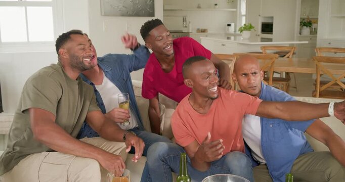 Diverse male friends laughing and passing smartphone on couch, center man gesturing for selfie