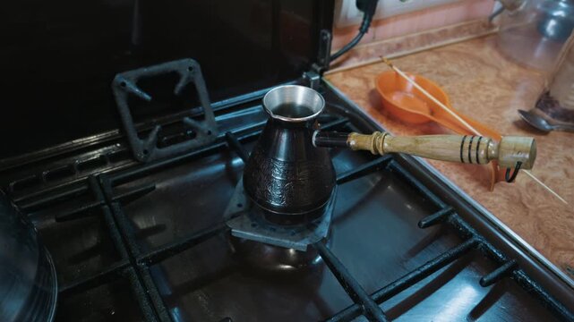 Metal coffee pot with a wooden handle sits on a gas stove burner for traditional brewing. The kitchen counter features an orange utensil rest. Traditional coffee brewing concept.