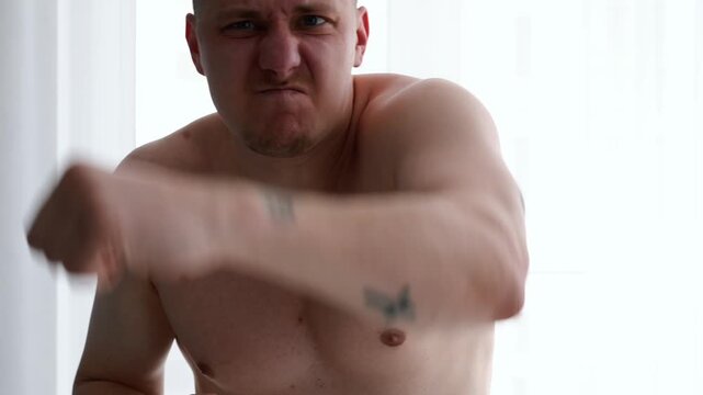 Muscular shirtless man in a bright room performing boxing-style punches with expressive hand movements, representing training and physical fitness.