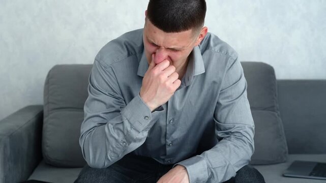 Young man in a gray shirt sitting in a bright room covering his nose with his hand, reacting to an unpleasant indoor odor and possible ventilation problem.