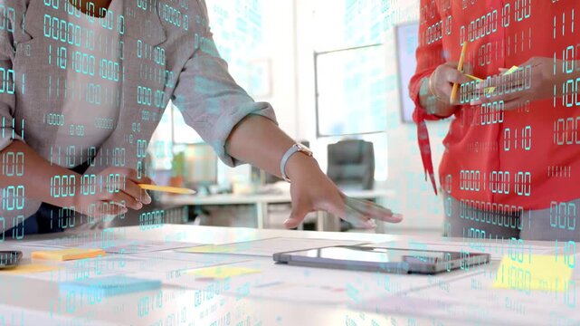 Left woman pointing other woman holding notes aligning design and tapping tablet under data overlay