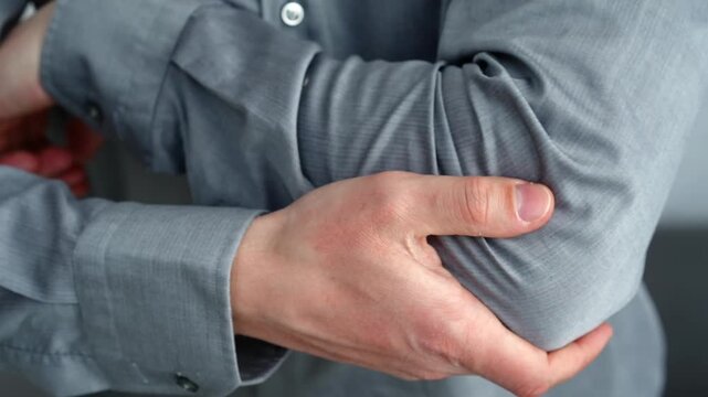 Young European man sitting in a bright room, holding his elbow and showing signs of pain after strain or overuse during physical activity or work.