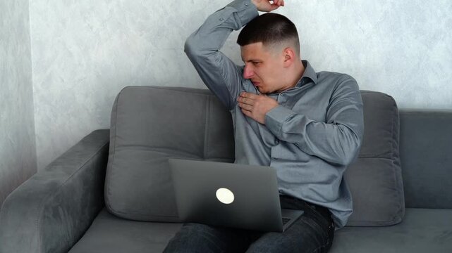 Man working remotely on a sofa in a warm room, feeling overheated and self-conscious due to sweating caused by hot weather while continuing work.