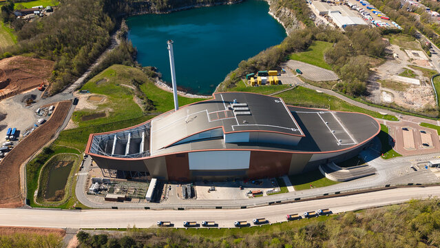 Aerial view of Encyclis Newhurst ERF featuring modern industrial architecture, a tall chimney, and a deep blue quarry lake under bright sunlight in Shepshed, England, United Kingdom.