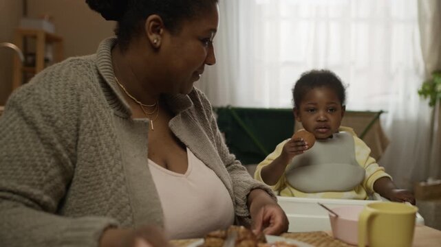 Zoom out shot of loving African American family members sharing modest meal together at home, with mother chatting with teenage daughter and toddler girl eating cookie in high chair