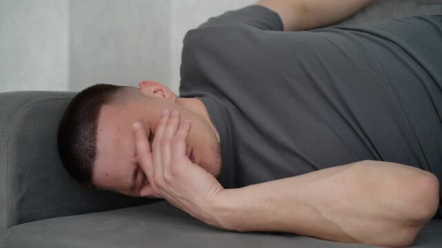 Brunette man lying on a gray sofa, waking up from a short rest and rubbing his face, expressing sleepiness and relaxation.