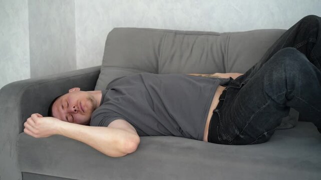 Brunette man lying on a gray sofa, waking up from a short rest and rubbing his face, expressing sleepiness and relaxation.
