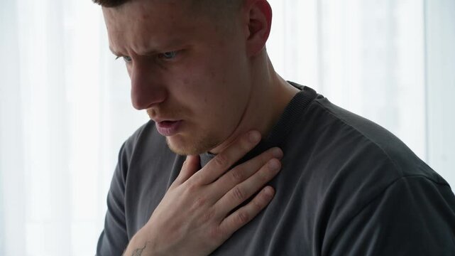 Young man in a gray t-shirt hiccuping in a bright room, reacting with discomfort after drinking water too quickly. Natural body reaction.