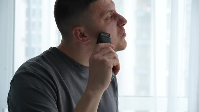 Young brunette man trimming his beard and mustache with an electric trimmer in a bright room, shaping facial hair as part of his daily grooming routine before going out.