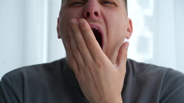 Close-up of a young man yawning in a bright room, showing signs of sleepiness in the morning. Concept of fatigue and waking up.