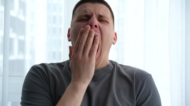Young man yawning in a bright bedroom, showing sleepiness and tired morning state. Concept of rest and daily routine.
