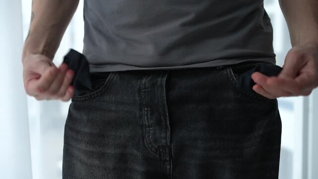 Young male showing empty pockets on black pants, expressing that he has no money left. Concept of financial shortage and economic stress