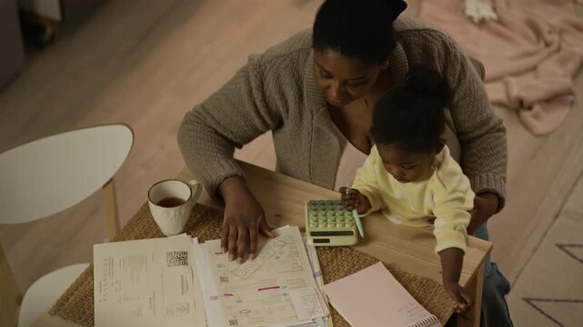 High angle shot of young Black mother sitting at table at home and reviewing overdue utility bills holding toddler daughter on laps, struggling with financial hardship while taking care of child