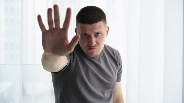 Man in a bright indoor room waving his hand in greeting but appearing visibly annoyed. Focus on forced politeness and emotional contradiction