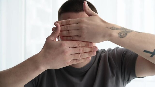 Young man in a bright room playfully covering and uncovering his face with his hands, rapidly changing facial expressions and mood. Concept of shifting emotions and playful behavior.