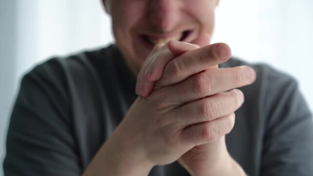 Man in a light room wearing a gray t-shirt making a sly facial expression while rubbing his hands together, symbolizing anticipation, strategy, or a clever idea forming