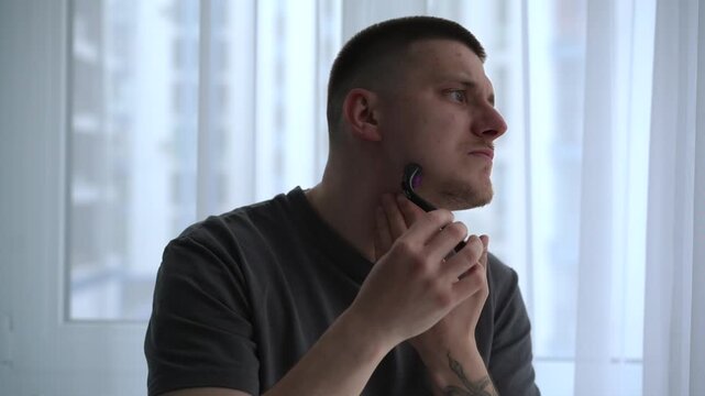 Young man using a dermaroller on his face as part of a beard growth routine, rolling the device over the skin to enhance absorption of minoxidil. 