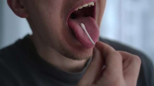 A casually dressed young man in a gray t-shirt swabs the inside of his mouth to collect saliva for analysis. The action suggests genetic testing, health screening, or laboratory sample collection