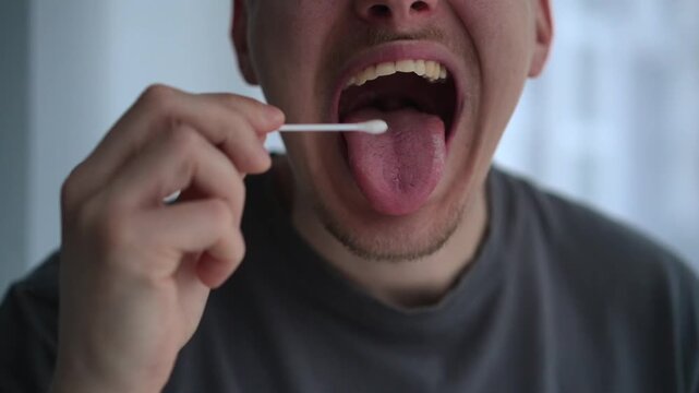 A young man takes a saliva sample with a cotton swab, placing it inside his mouth to gather material for testing. The scene reflects a simple at-home procedure for DNA testing or medical diagnostics.