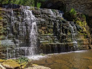 A tiered limestone waterfall cascades over moss-covered rock ledges into a shallow pool during a sunny day in a summer forest. © Matt