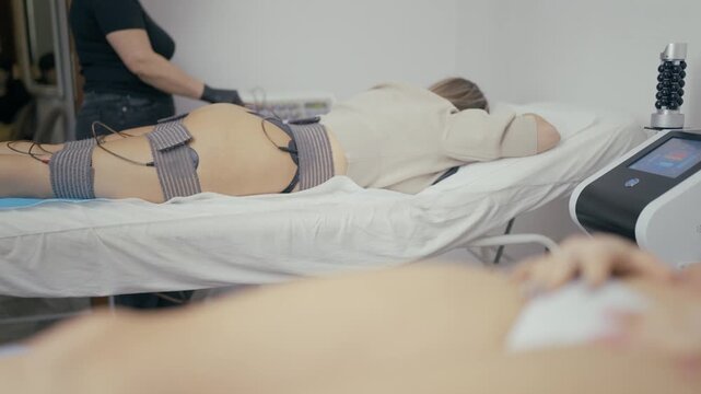 A woman undergoes an electro stimulation treatment at a beauty salon to improve body contour and muscle tone. She lies on a couch receiving an aesthetic therapy session for slimming and firming.