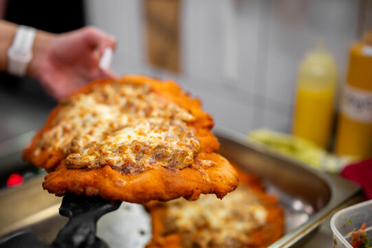 A golden-brown deep-fried Hungarian langos being lifted with a spatula, topped with melted cheese and savory meat, against a blurred kitchen background with squeeze bottles.