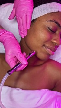 A woman enjoys a facial treatment as a beauty professional carefully performs dermaplaning on her skin. The setting is a beauty salon with bright lighting.