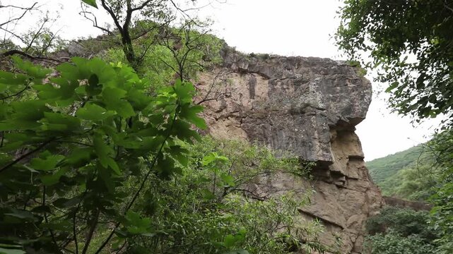 The video begins with a close-up of a highly textured porous rock overhang. The camera then slowly tilts upwards revealing a steep, rugged cliff face with green vegetation.

