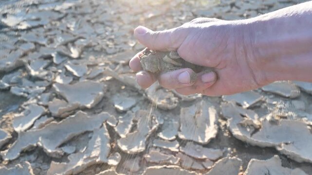 hand holding dry, arid soil that crumbles into fine dust as it is rubbed between the fingers. In the background, a barren and parched landscape stretches out, emphasizing drought conditions