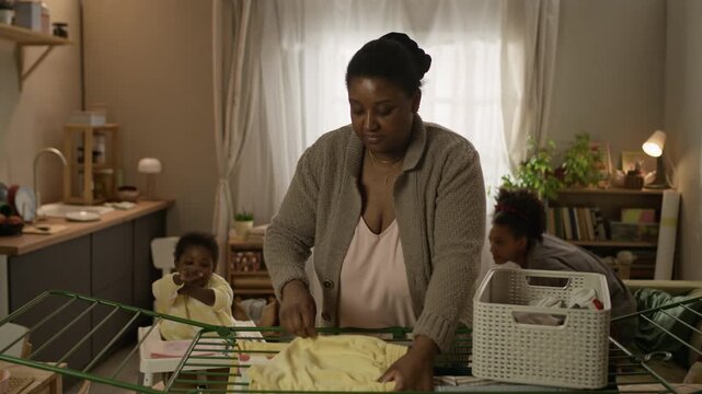 Medium long shot of teenage Black girl playing with her little sister sitting in high chair, while their mother hanging laundry on drying rack in foreground, sharing small modest apartment together