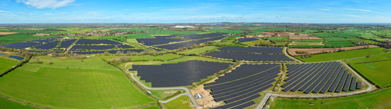 Aerial view of a vast solar farm with rows of photovoltaic panels across green fields under a blue sky in Nailstone, England, United Kingdom.