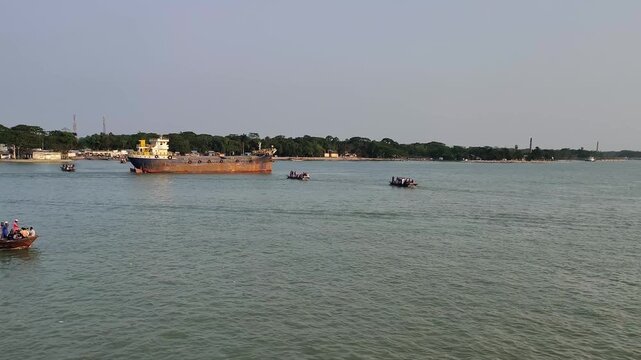 4K Cinematic wide view of large industrial cargo ship and traditional passenger boats navigating a busy river in kirtonkhola river,Barishal, Bangladesh.
