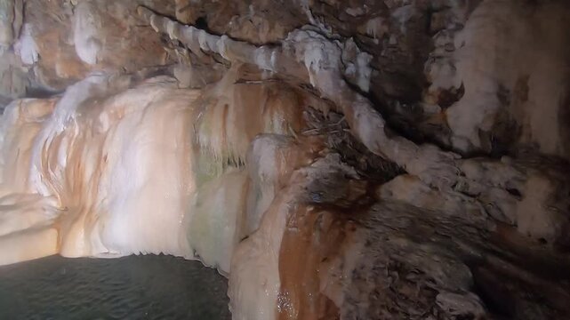 A panning shot reveals gentle water flowing over smooth, rounded rock formations inside a dark cave, some with green moss.
