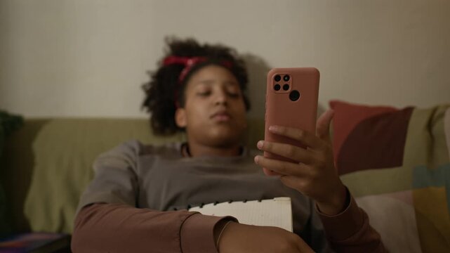 Selective focus shot of teenage Black girl lying on couch at home holding notebook and scrolling newsfeed on smartphone, absorbed into screen with apathetic expression