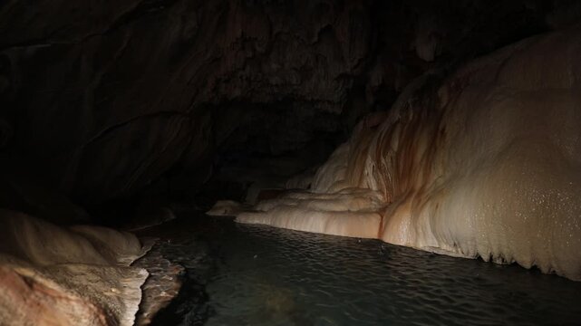 A panning shot reveals gentle water flowing over smooth, rounded rock formations inside a dark cave, some with green moss.
