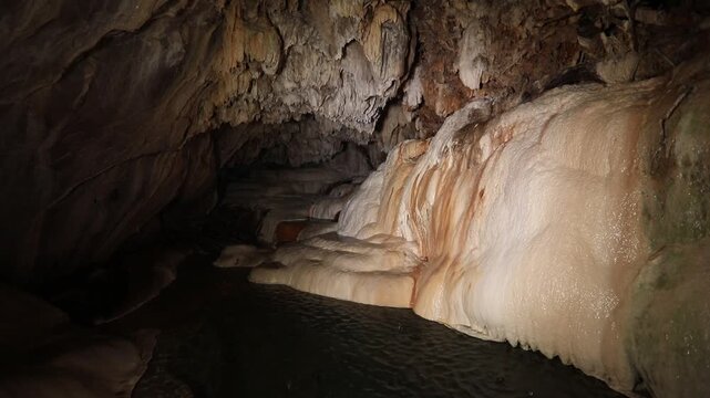 A panning shot reveals gentle water flowing over smooth, rounded rock formations inside a dark cave, some with green moss.
