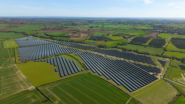 Aerial view of a large solar farm with rows of photovoltaic panels set amidst green agricultural fields and hedgerows under a clear blue sky Nailstone, England, United Kingdom.