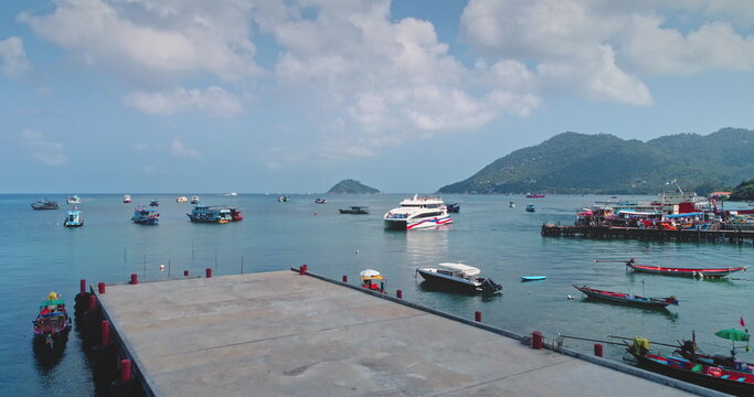 Aerial view of ferry approaching Koh Samui Island pier in Thailand, with traditional long tail boats and other vessels sailing in a beautiful tropical bay under a cloudy sky