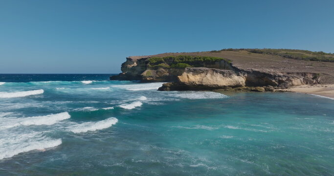 Penisok, Lombok, Indonesia: Ocean waves breaking on the sandy beach with rugged coastline cliff meeting the blue sky, creating a serene and natural tropical landscape. Wild nature. Aerial drone fligh