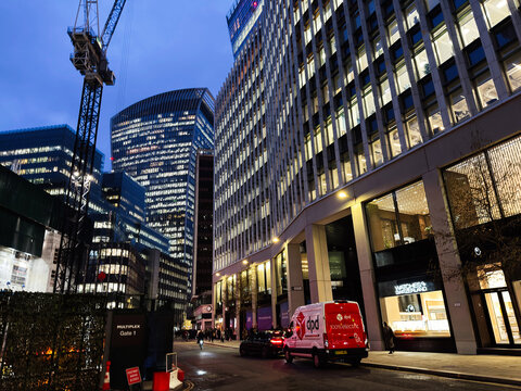 Evening business streetscape in London, UK with illuminated office buildings, delivery van and construction crane among modern skyscrapers