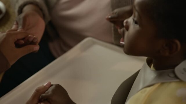Close up shot of African American toddler girl wearing bib sitting in high chair and holding hands of unrecognizable family members, praying before having dinner at home