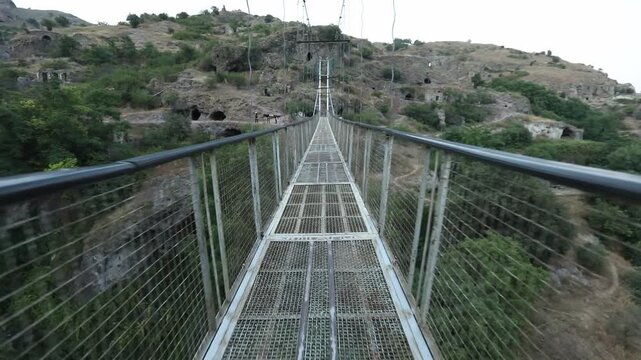 First person view walking across a long metal suspension bridge spanning a deep lush gorge leading towards ancient cave dwellings carved into the rocky mountainside in the distance.

