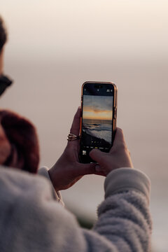 Hands holding phone capturing ocean sunset in natural photo style on soft sky background, concept of travel memories