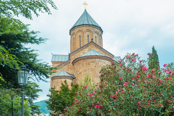 Metekhi Church in Tbilisi old town surrounded by greenery and flowers in the foreground. Historic Georgian religious architecture blending with nature and urban environment. Travel destination © samael334
